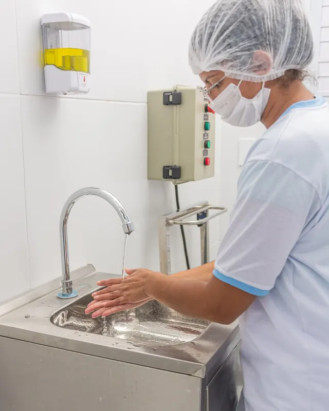 Funcionária de touca e máscara lavando as mãos na pia de inox